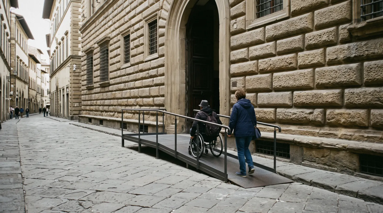 A metal ramp beside a stone doorway in a Florentine palace facade, a visitor in a wheelchair with a companion approaching, step-free access in Florence