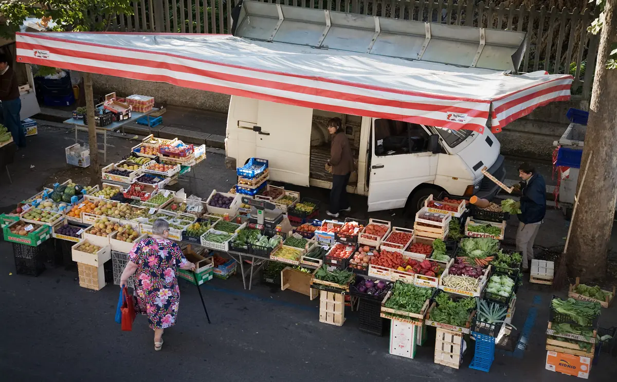 A Florence street market stall with fruit and vegetable crates, each carrying a handwritten price tag